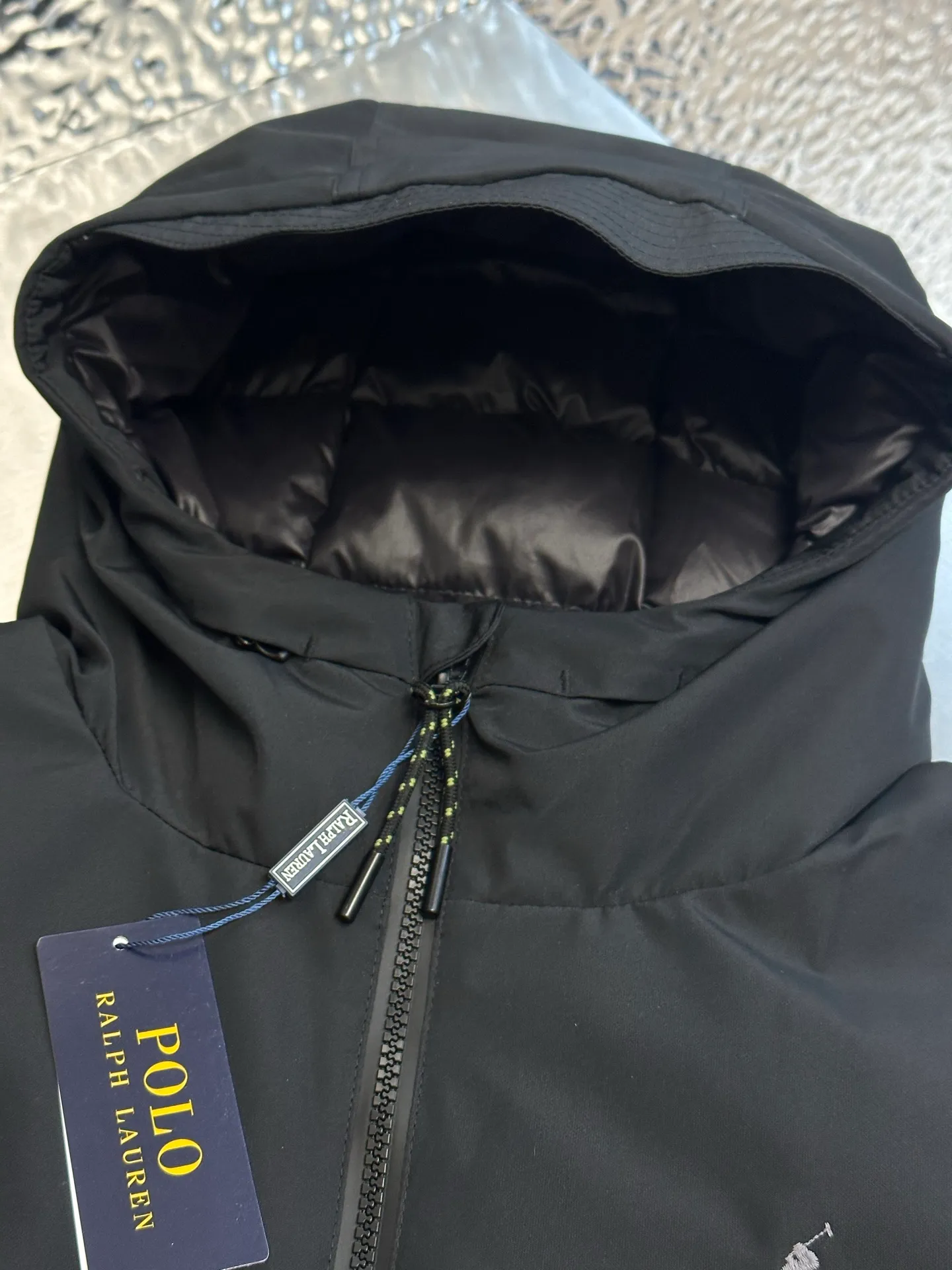 P970 Ralph Lauren logo hooded down jacket. The density of high-density nylon taffeta is about 210T-300T. The surface is smooth and slightly matte. It has excellent windproof and waterproof performance (with DWR water-repellent treatment). It has strong wrinkle resistance and is easy to care for. It is in line with the positioning of American casual "durable and practical".  The inner lining is made of 100% polyamide. The high-density and tight weaving process prevents fine lint from getting out of the gaps in the fabric. The smooth surface improves the putting on and taking off experience, and the excellent wear and tear resistance ensures damage caused by frequent use, while the light and soft texture does not affect the overall weight.  It uses 90 white duck down with a weight of 220g to ensure excellent thermal insulation performance. After strict screening and processing, the velvet is not easily drilled, ensuring durability. The classic jacket has a slightly loose fit, with slightly dropped shoulder lines (the shoulder width is 1-2cm wider than the standard fit). The length reaches the hips, and the sleeves are slightly tapered straight sleeves, taking into account freedom of movement and casual style.  The adjustable Velcro on the cuffs allows you to freely adjust the tightness according to the thickness of your wrist and personal preferences, effectively preventing cold wind from intruding, and improving the warmth performance. Two zippered pockets on the front are convenient for placing frequently used items, and the zipper design ensures that items are not easily lost. A built-in pocket on the left side of the chest provides a safe storage space for some small private items.  The drawstring design at the hem allows you to adjust the shape of the jacket according to your body shape and dressing needs. Whether it is loose and casual or slim and neat, you can easily control it.  Polo horse logo on the left chest: flat satin stitch embroidery (satin embroidery), using 100% polyester high color fastness embroidery thread (the color number is consistent with the brand's standard red color card), the stitch density is 12-15 stitches/cm, the outline of the horse logo is clear, and the details of the rider and horse are burr-free. Precise positioning through the template (12cm from the bottom of the collar and 8cm from the placket) ensures symmetry and centering.  size：s m l xl